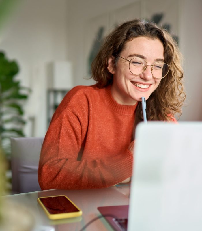 Happy young woman using laptop sitting at desk writing notes while watching webinar, studying online, looking at pc screen learning web classes or having virtual call meeting remote working from home.